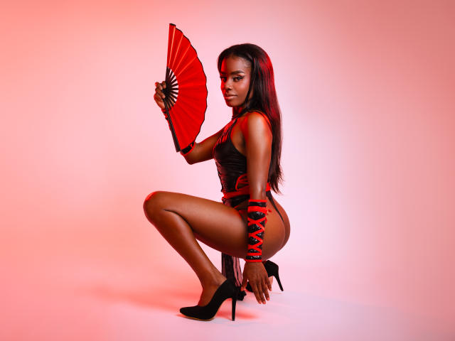 MiaHopson, squatting, holding red fan, wearing black latex lingerie, red wrist cuffs, black high heels, with dark brown skin and long red-black hair, showing her ass.
