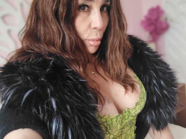 IssaMilfX with wavy brown hair, green lace top, and black fur coat, showing her cleavage, wearing a necklace, with a slightly pouty expression, in a pink room.