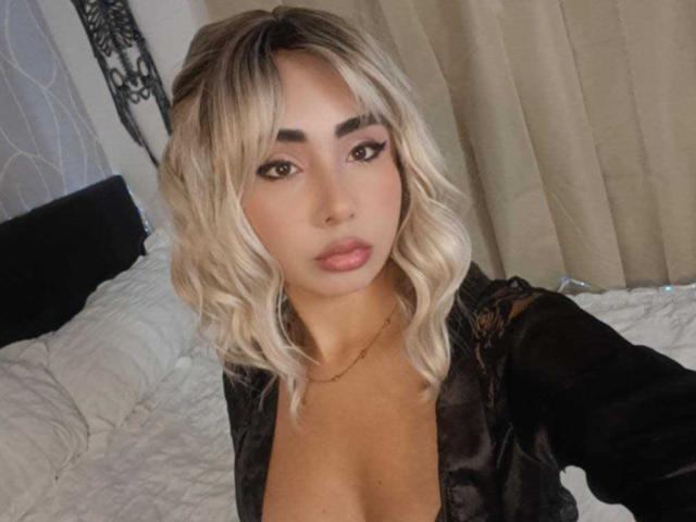 EmilyValk, con i capelli biondi e ondulati, indossa una camicetta nera in pizzo che mette in mostra il décolleté, ha labbra piene ed è rivolta verso la fotocamera con un trucco occhi scuro e allungato..