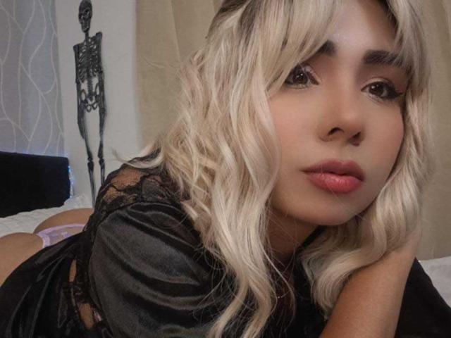 EmilyValk with blonde wavy hair, wearing a black lace top, lying on a bed, looking at the camera with pink lips, showing her cute ass, in a sexy pose.