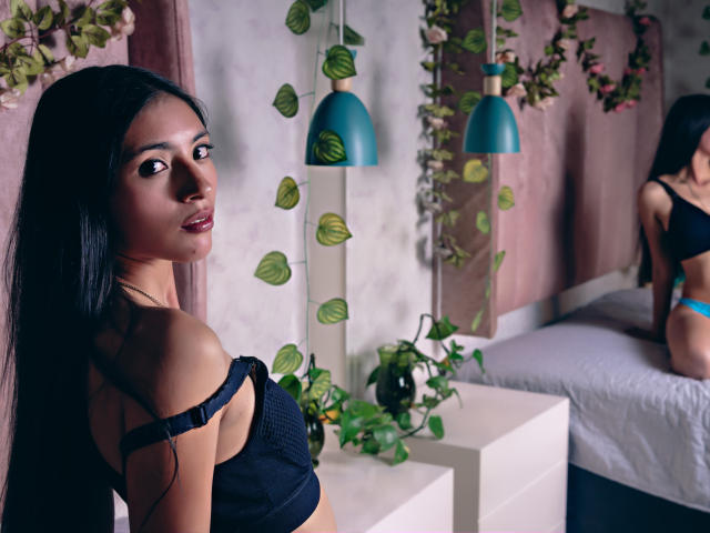 Alana Moore met lang zwart haar, donkere ogen en een gemiddelde borst, gekleed in een zwarte bh, kijkt om, staand bij een witte nachtkastje met groene planten en blauwe hanglampen..