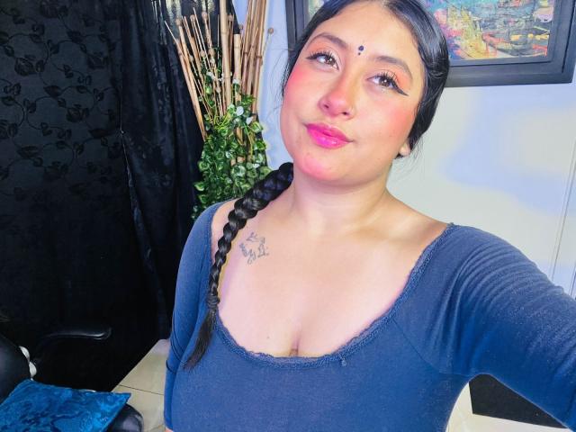 KalpanaAisha with dark hair in a braid, pink lipstick, and a small tattoo on her chest, wearing a blue top, showing her boobs, smiling slightly, in a room.