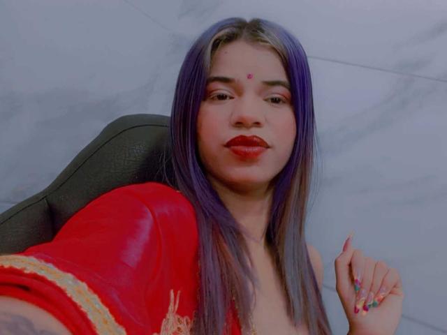 KairaDayamai with purple and blonde hair, red lipstick, red dress, small red bindi on forehead, holding up right hand with colorful nails, seated in gray chair.
