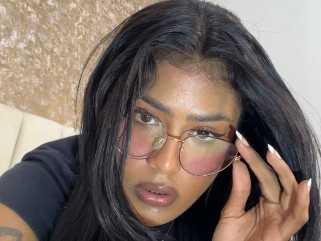 Babilett, avec ses longs cheveux noirs, portant des lunettes aux verres légèrement rosés, touchant son visage avec ses doigts manucurés de vernis blanc, a des lèvres roses et brillantes, et porte une chemise noire..