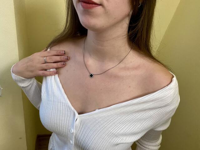 Nesie with fair skin, brown hair, and pink lipstick, wearing a white ribbed cardigan, exposing her tits, hand on chest, silver necklace, pink nails, and a ring.
