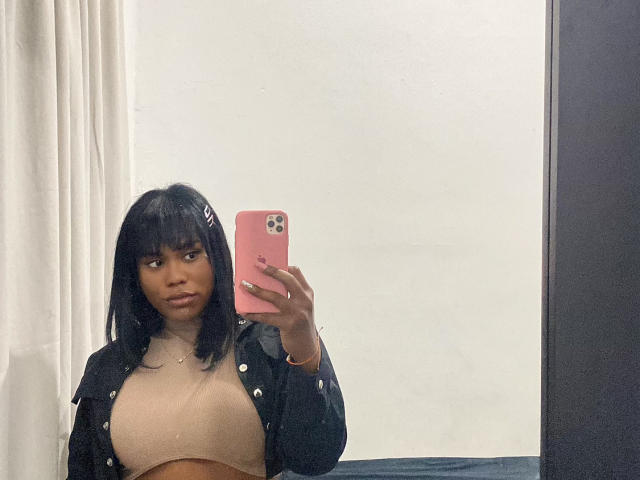 AshleyRob with dark skin, black bob, pink phone, black jacket, beige top, holding phone in mirror, showing her tits, looking at camera, white curtain on left.