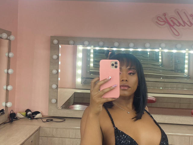 AshleyRob with medium brown skin, black bob, pink eyeshadow, holding pink phone, showing small breasts in black strappy top, mirror with lights, nail polish, and necklace.