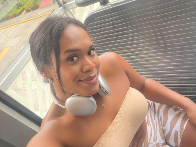 AshleyRob with dark skin, smiling, wearing a beige strapless top, white zebra-patterned skirt, gray headphones, black hair in a ponytail, sitting in a car.