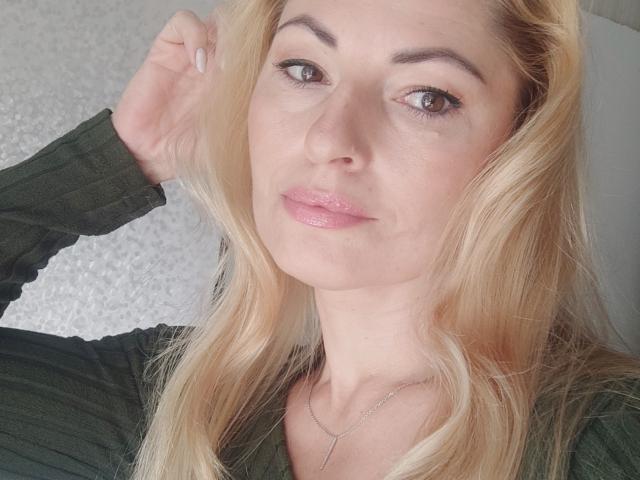 Sienna Taylor, com pele bronzeada, cabelos longos e pretos e um colar de ouro, usando luvas pretas, cobrindo seus seios grandes e brilhantes com as mãos. Fundo de veludo vermelho..