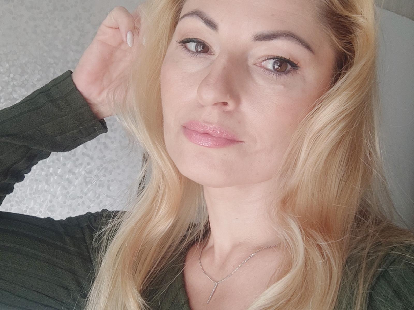 Sienna Taylor, con la pelle abbronzata, i lunghi capelli neri e una collana d'oro, indossa dei guanti neri, coprendo con le mani i suoi seni grandi, lucidi e scoperti. Sfondo in velluto rosso..