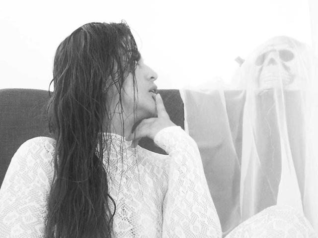 SkylarDesire with wet dark hair, wearing a white lace top, sitting on a dark couch, looking slightly to the side with a subtle, seductive expression. PMS tits, small earrings.