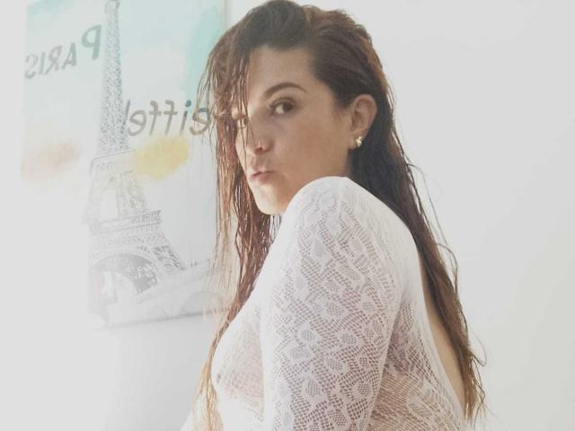 SkylarDesire with wet brown hair, wearing a white, see-through, lace-up back top, looking over her shoulder, showing her back and small, round ass.