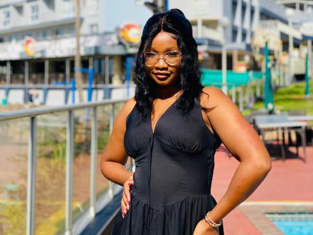 A photo of SugarSpiceQueen with dark skin, black wavy hair, glasses, and a black dress, standing with hands on hips, showing her tits and ass.