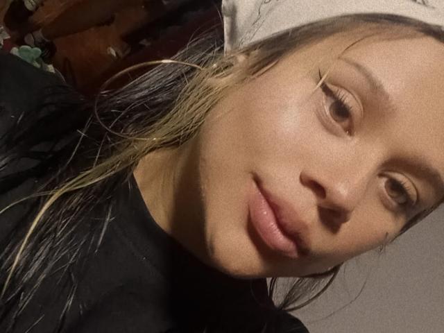 SamantaGreen has a slightly sweaty face, blonde and brown hair, and is wearing a beige hat, with black clothes and subtle makeup, looking at the camera with a slight smile.