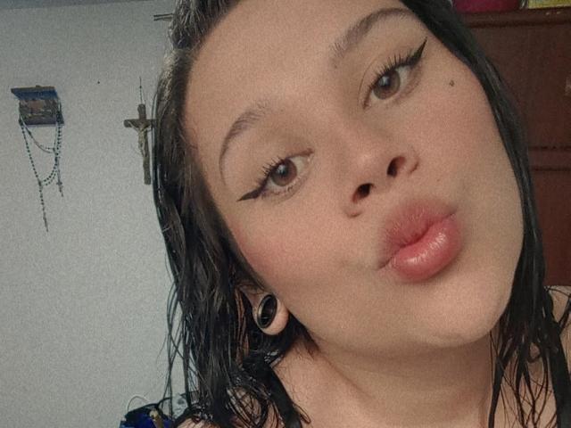 SamantaGreen with wet black hair, thick black eyeliner, and pouty pink lips, wearing black ear gauges, looking up with brown eyes. Soft, light-skinned face, slightly tilted.