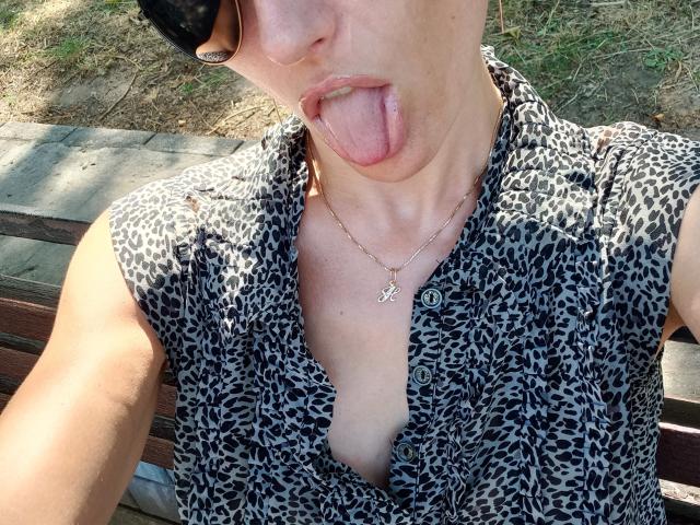 BellaJulls, met haar tong uit, draagt een shirt met luipaardprint, laat een beetje decolleté zien, en een kleine gouden ketting met een 'P'-bedel, met zonlicht op haar gezicht..