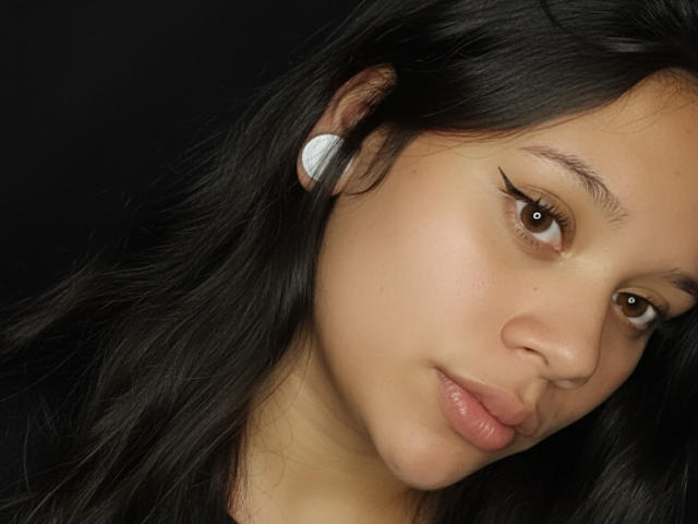 SamantaGreen has tan skin, dark black hair, brown eyes, and wears white earbuds. Her full lips are slightly pouty, and she's looking down with a subtle smile.