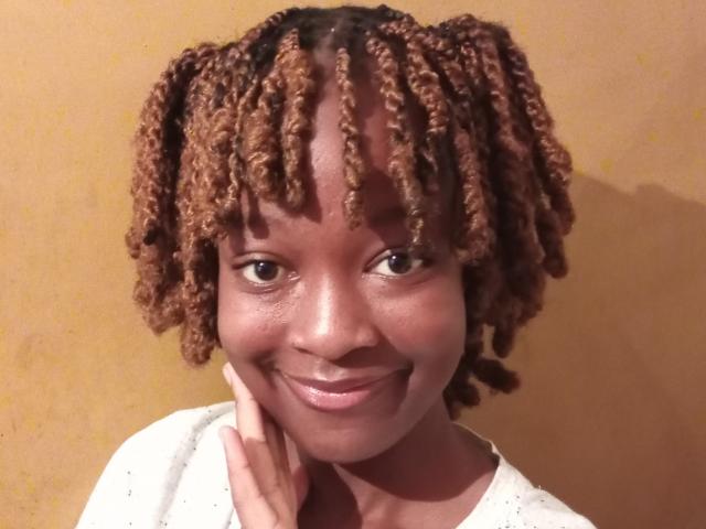 NataWilma with brown, curly, shoulder-length dreadlocks, smiling, brown eyes, dark skin, white shirt, hand touching cheek, yellow background, slightly glossy lips, relaxed and happy expression.
