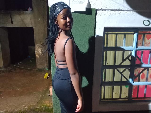 NataWilma with dark skin, black braided hair, wearing a black, backless, high-waisted dress, showing her ass, standing against a green wall with a colorful window.
