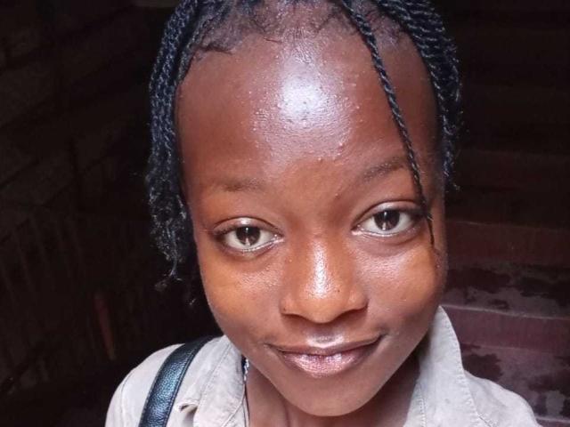 NataWilma's shiny brown face with braided black hair, brown eyes, and subtle smile, wearing a beige shirt, with a blue strap visible over her shoulder.