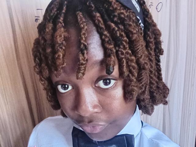 NataWilma has dark brown, curly, medium-length afro hair with black and brown tips, wearing a blue bowtie, white shirt, and black headband, looking straight at the camera.