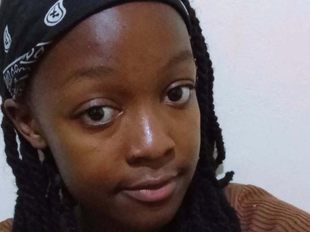 NataWilma's dark brown face with shiny skin, black dreadlocks, and a black bandana, wearing a brown, ribbed shirt, looking slightly to the side with a subtle smile.