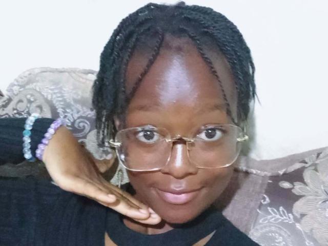 NataWilma with dark skin, wearing clear glasses, black top, purple and blue bead bracelet, and braided hair, has a hand near her chin, smiling softly.