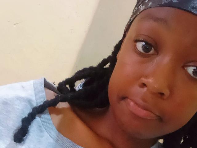 NataWilma with dark brown skin, black dreadlocks, wearing a gray shirt, and a black headscarf, looking slightly to the side with a subtle smile.