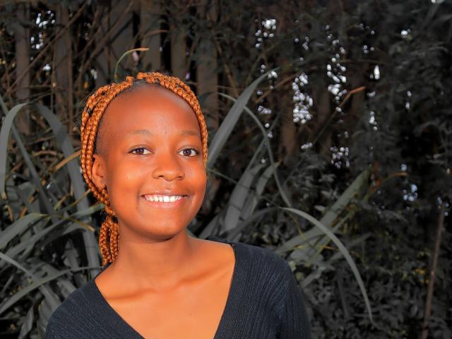 NataWilma with brown braids, smiling, wearing a black V-neck, has a cute ass, and is looking to the side with bright eyes in a sunny outdoor setting.