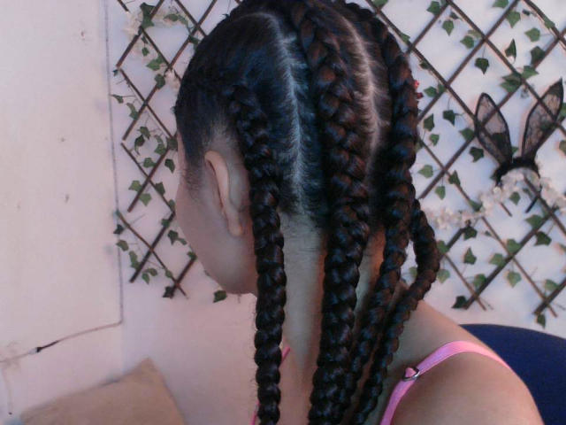 SexLaddy with long, dark braided ponytails, side profile, wearing pink bra straps, against white wall with vine pattern, showing smooth brown skin, subtle makeup.