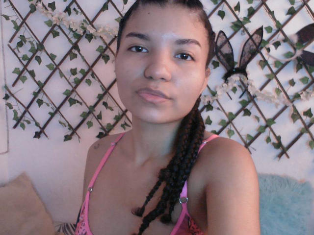 A selfie of SexLaddy with medium brown skin, long braided black hair, pink bra, small tits, and a neutral expression, standing in front of a vine-covered lattice.