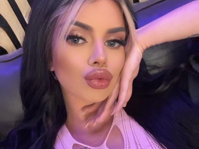 IvyRaven with pouty pink lips, blonde and black hair, heavy eyeliner, and a hand touching her cheek, wearing a white top with cutouts, sitting on a black leather seat.