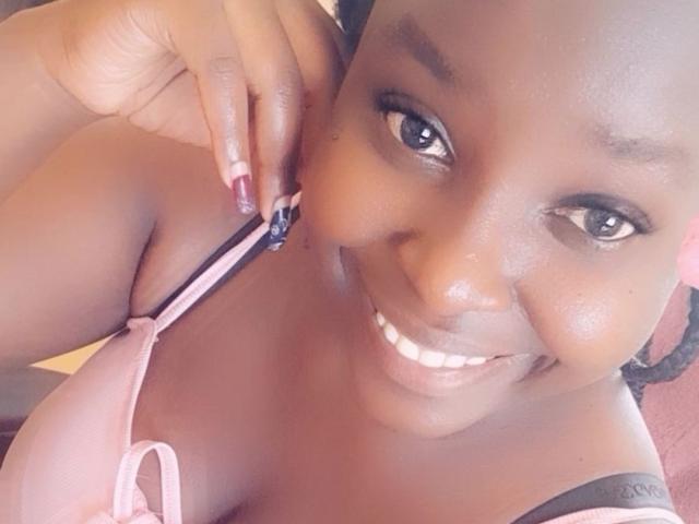 CasieStar with dark skin, smiling, pulling pink bra strap with purple nail, showing cleavage, black bra strap, and shiny lips, in a selfie.