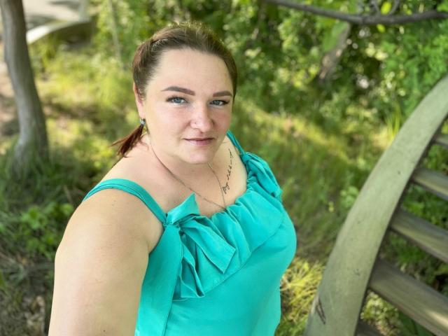 AngelQSky with medium tits, wearing a teal dress with ruffles, standing outdoors, brown hair in a ponytail, green earrings, and a necklace, smiling at the camera.