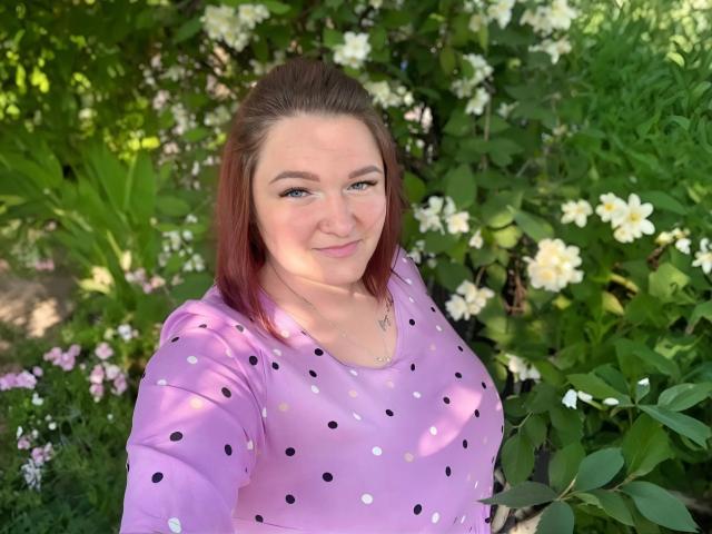 AngelQSky with red hair, purple polka dot shirt, smiling, tits visible, green eyes, fair skin, standing in garden with white and pink flowers.