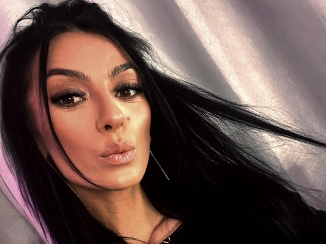 KinkyAmelie mit langen, schwarzen Haaren, voller Lippen und dunkelbraunen Augen, die einen Kuss macht, mit dezentem Make-up und Reifenohrringen, in einem leicht unscharfen Selfie..