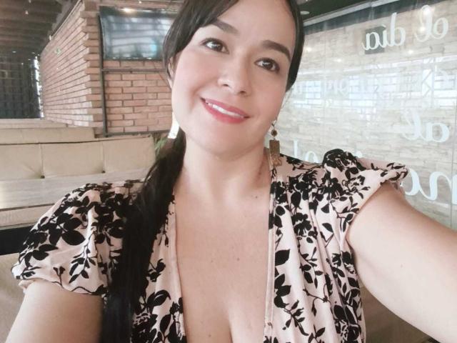 TamaraFoxy con un vestido floral negro y rosa con volumen, que muestra el escote, sonriendo, ojos marrones, cabello negro en una coleta, piel clara, sentada en una cafetería, con luces brillantes en el fondo..