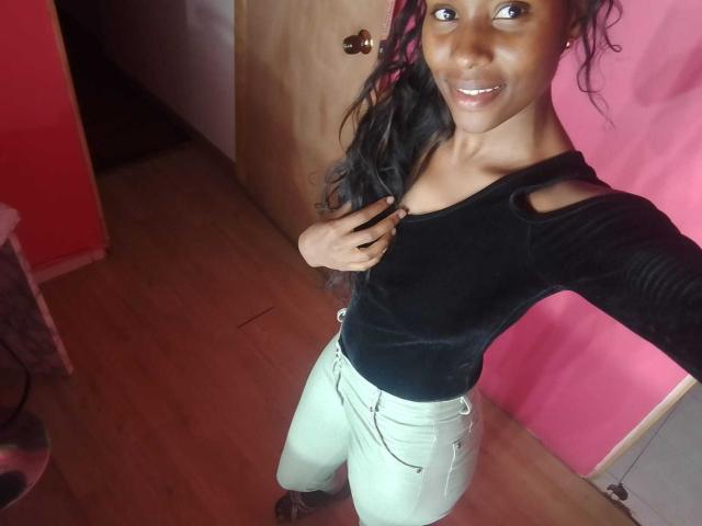 HornyCocoa with dark skin, long black hair, black velvet top, cutout shoulder, light green pants, smiling, standing in pink-walled room, hand on chest.