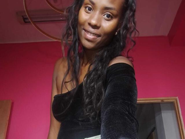 HornyCocoa with dark curly hair, smiling, wearing a black off-shoulder velvet dress, standing in a room with a red wall and a circular ceiling light.