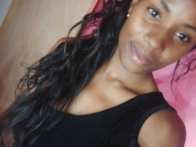 HornyCocoa with dark skin, long wavy black hair, small earrings, smiling, black off-shoulder top, showing bare shoulders, looking at camera, pink background.
