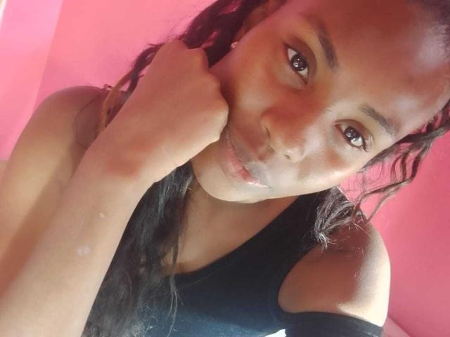 HornyCocoa with dark brown skin, wavy black hair, and brown eyes, resting her cheek on her hand, wearing a black tank top, smiling softly. Pink background.
