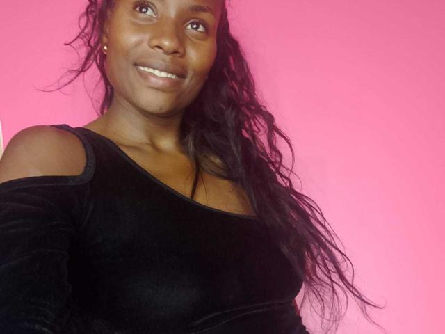 HornyCocoa with dark skin, wavy black hair, smiling, wearing a black off-shoulder top, showing her tits, against a pink background.