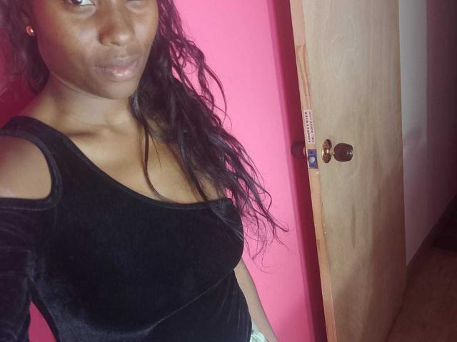 HornyCocoa with dark brown skin, long wavy black hair, and shiny lips wearing a black velvet top with cutout shoulders, standing in front of a wooden door.