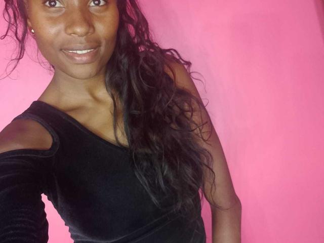 HornyCocoa with dark skin, long wavy black hair, smiling, wearing a black velvet off-shoulder top, pink background, visible shoulder and upper chest, small earrings.