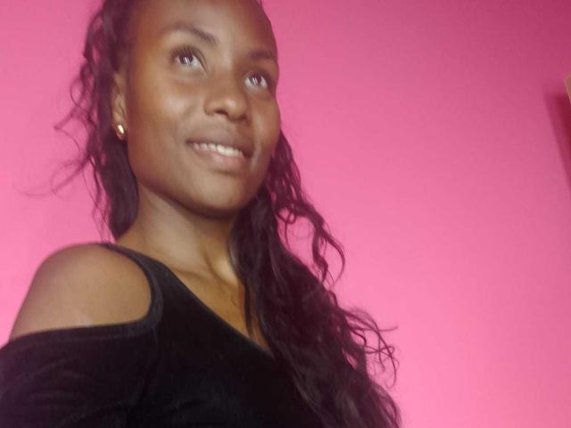 HornyCocoa with dark brown skin, long wavy black hair, smiling, wearing a black off-shoulder top, small gold earrings, pink background.
