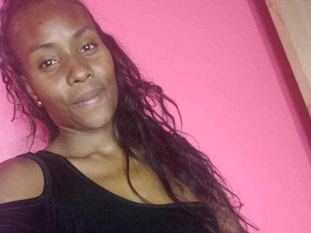 HornyCocoa with dark skin, long wavy black hair, and small earrings, wearing a black top, smiling softly at the camera. Pink background.