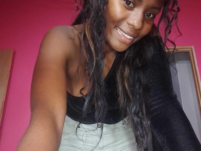 HornyCocoa with dark skin and curly black hair, smiling, wearing a black tank top and light green pants, showing her bare shoulders and chest. Her tits are small.