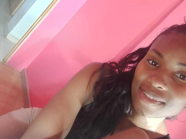 HornyCocoa with long black hair, smiling at the camera, wearing a black top, pink wall in background, wooden floor, and white pants, taking a selfie.