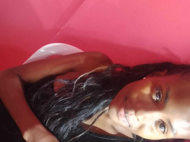 HornyCocoa with dark brown skin, long black hair, and brown eyes, wearing a black top, lying down with a white balloon, smiling subtly. Red background.
