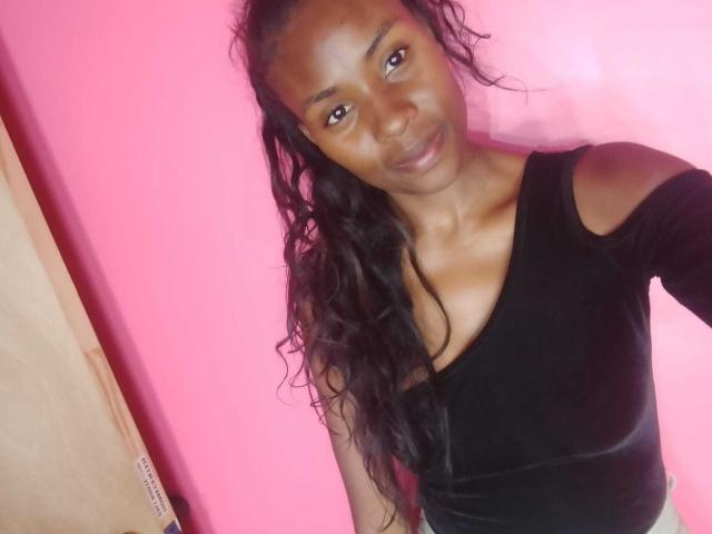 HornyCocoa with dark brown skin, long wavy black hair, small boobs, wearing a black V-neck top, smiling, standing against a pink wall, and a wooden door on the left.